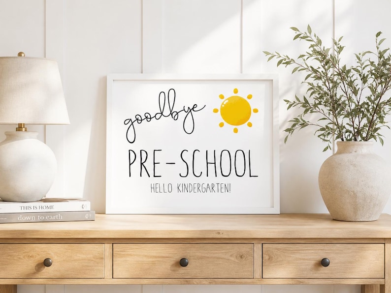 Puede incluir: Un cartel enmarcado con las palabras "goodbye PRE-SCHOOL HELLO KINDERGARTEN!" en negro. Un sol amarillo est&aacute; a la derecha de la palabra "goodbye". El cartel est&aacute; sobre una mesa de madera, con una l&aacute;mpara y un jarr&oacute;n.
