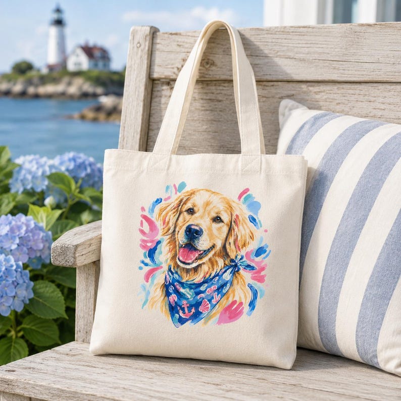 May include: A cream-colored tote bag featuring a watercolor illustration of a golden retriever wearing a blue bandana with an anchor design. The dog is surrounded by pink and blue brushstrokes. The bag has cream-colored handles.