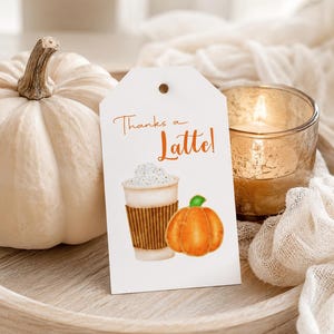 May include: A white pumpkin and a tag with the words "Thanks a Latte!" and a watercolor illustration of a latte and a pumpkin. A lit candle in a glass holder is also visible.