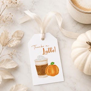 May include: A white gift tag with the text "Thanks a Latte!" features a watercolor illustration of a coffee cup and a pumpkin. A beige ribbon is attached to the tag. A cup of coffee and a white pumpkin are also in the image.