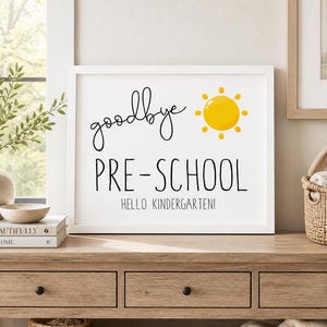 Puede incluir: Una impresi&oacute;n blanca enmarcada con las palabras "goodbye PRE-SCHOOL HELLO KINDERGARTEN!" en escritura negra y letras de bloque, con una ilustraci&oacute;n de sol amarillo. La impresi&oacute;n se muestra en una mesa de madera con un jarr&oacute;n de vegetaci&oacute;n y una cesta decorativa.