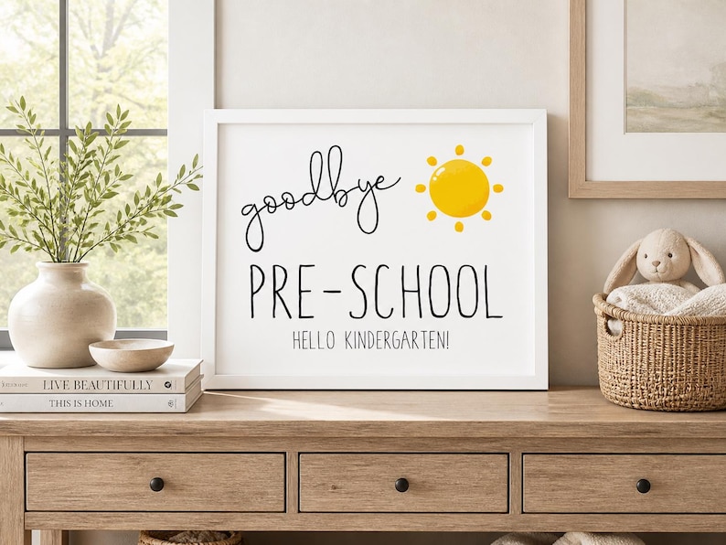 Puede incluir: Una impresi&oacute;n blanca enmarcada con las palabras "goodbye PRE-SCHOOL HELLO KINDERGARTEN!" en escritura negra y letras de bloque, con una ilustraci&oacute;n de sol amarillo. La impresi&oacute;n se muestra en una mesa de madera con un jarr&oacute;n de vegetaci&oacute;n y una cesta decorativa.