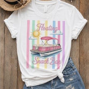 Może przedstawiać: Biała koszulka z kolorową grafiką różowej łodzi pontonowej z napisem "Floating Snack Bar". Koszulka jest połączona z dżinsowymi szortami i słomkowym kapeluszem.