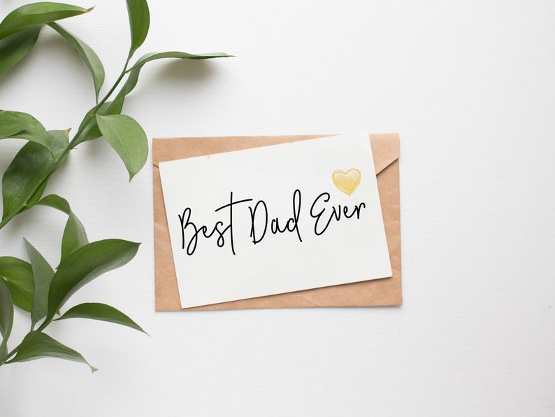 May include: A greeting card with the text "Best Dad Ever" in black script, a yellow heart, and a kraft paper envelope. Green leaves are in the upper left corner. The card is on a white background.