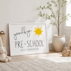 Puede incluir: Un letrero blanco enmarcado con las palabras "goodbye PRE-SCHOOL" y "HELLO KINDERGARTEN" en escritura negra. Un sol amarillo est&aacute; a la derecha de "goodbye". El letrero est&aacute; sobre una alfombra clara.