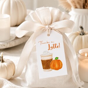 May include: A beige fabric gift bag tied with a cream ribbon, with a tag that reads "Thanks a Latte!" and features a watercolor illustration of a latte and a pumpkin. White pumpkins and candles are in the background.