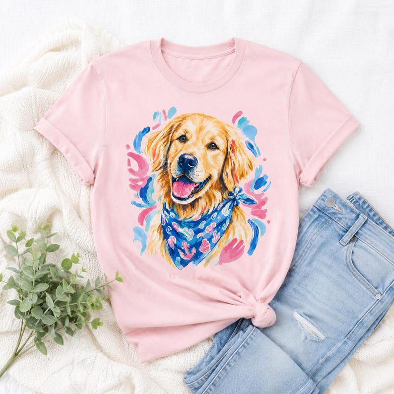 May include: A light pink t-shirt featuring a watercolor illustration of a Golden Retriever dog wearing a blue bandana with anchor designs. The dog has a happy expression. The shirt is paired with light blue denim jeans.