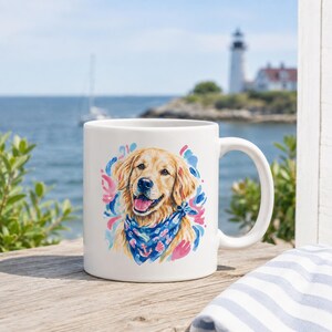 May include: White ceramic mug featuring a watercolor illustration of a Golden Retriever wearing a blue bandana with anchor and seashell designs. The dog is surrounded by pink and blue brushstrokes. The mug has a white handle.