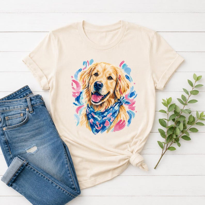 May include: Cream-colored t-shirt featuring a watercolor illustration of a Golden Retriever wearing a blue bandana with anchor and seashell designs. The dog is surrounded by blue, pink, and purple brushstrokes. The shirt is paired with blue denim jeans.