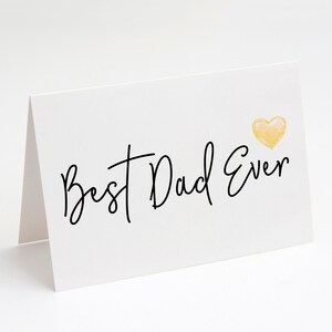 May include: A white greeting card with the text "Best Dad Ever" in black script. A small, yellow watercolor heart is in the upper right corner. The card is folded and standing upright.