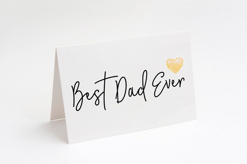 May include: A white greeting card with the text "Best Dad Ever" in black script. A small, yellow watercolor heart is in the upper right corner. The card is folded and standing upright.