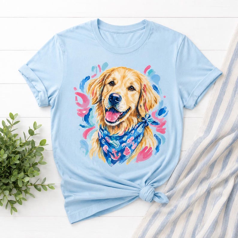 May include: Light blue t-shirt featuring a smiling golden retriever wearing a blue bandana with anchor and seashell designs. The dog is surrounded by colorful brushstrokes of pink, blue, and green. The shirt is knotted at the bottom.