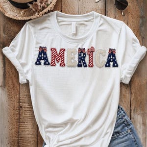 Könnte beinhalten: Weißes T-Shirt mit dem Wort "AMERICA" in rot, weiß und blau. Die Buchstaben sind mit Schleifen und patriotischen Mustern verziert. Das Shirt wird mit Jeansshorts und einem Strohhut kombiniert, was einen lässigen, sommerlichen Stil ergibt.