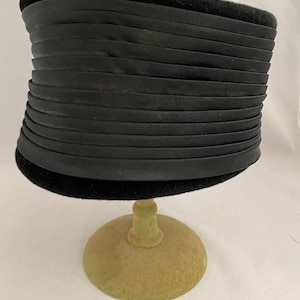 May include: A black velvet hat with a pleated band. The hat is displayed on a green stand.