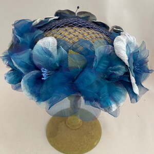 May include: A blue and white hat with a net and flower embellishments. The hat is on a stand.