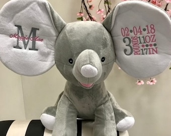 Op de afbeelding: Grijze pluche olifant knuffel met witte oren. De oren zijn geborduurd met de naam "Maggie Mae" en de datum "02-04-18", het gewicht "3 pond 11 oz" en de lengte "17 inch".