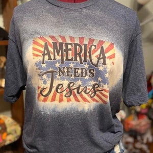 May include: A blue t-shirt with a distressed American flag design and the text "America Needs Jesus".