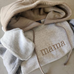 May include: A gray and beige hooded sweatshirt with the word "mama" embroidered on the front in a beige color.