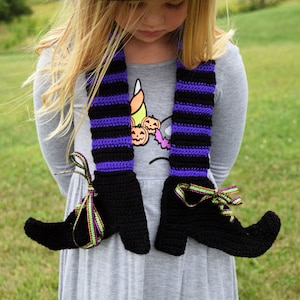 May include: A young girl wearing a black witch hat with a green ribbon and a black and purple striped crocheted scarf with witch boot ends. The scarf has green and gold ribbon accents.
