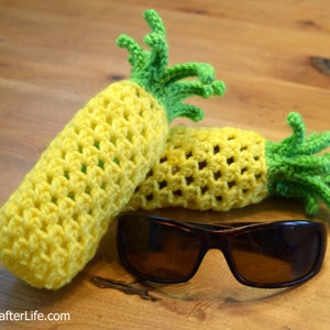 May include: Two crocheted pineapple-shaped holders for sunglasses, one yellow and one green, with a pair of brown sunglasses on a wooden surface.
