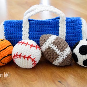 Puede incluir: Una bolsa de deporte de ganchillo azul y blanco con un asa blanca, acompañada de balones deportivos de ganchillo: un balón de baloncesto, béisbol, fútbol americano y fútbol. El baloncesto es naranja con líneas negras.