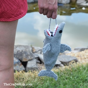 May include: A gray crocheted shark with a white and red open mouth. The shark is hanging from a string and is being held by a person's hand.
