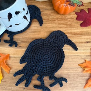 Puede incluir: Un posavasos de ganchillo azul marino con forma de cuervo, con alas y patas detalladas, está sobre una superficie de madera. También se ve una taza blanca con siluetas de pájaros negros y una pequeña calabaza naranja. El texto "The Crafter Life" está en la parte inferior.