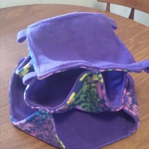 May include: A multi-level pet bed made of purple fleece with a colorful patterned fabric lining the inside. The bed is designed for small animals like hamsters or gerbils.