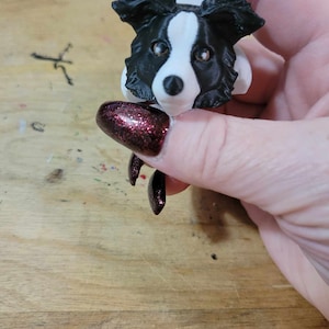 May include: A black and white border collie dog figurine with brown eyes. The figurine is being held in a hand with dark red nail polish.