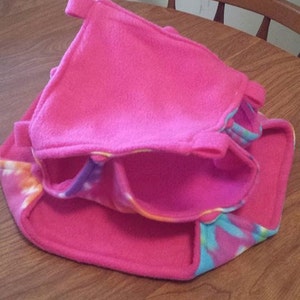 May include: A pink and blue fleece hamster hideout with a tie-dye pattern. The hideout has a flap that can be closed to create a cozy space for a small pet.