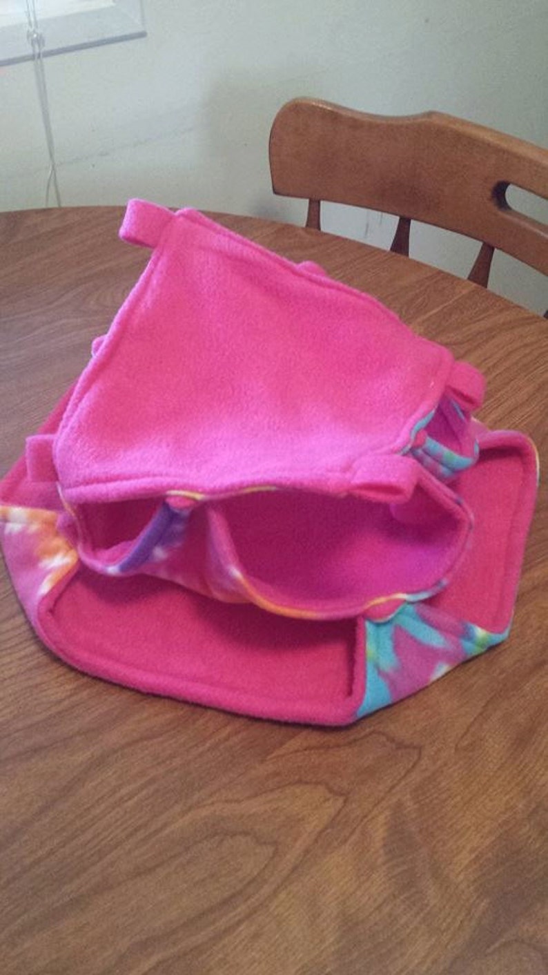 May include: A pink and blue fleece hamster hideout with a tie-dye pattern. The hideout has a flap that can be closed to create a cozy space for a small pet.