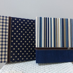 May include: Two fabric-covered photo albums with different patterns. One album has a blue and white gingham pattern with a brown ribbon. The other album has a blue background with white stars and a brown ribbon. Both albums have a dark blue bottom section.
