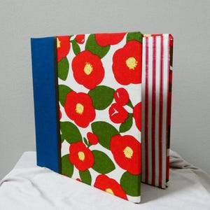 May include: Three decorative books with unique covers. One has a blue spine and a floral pattern of red flowers and green leaves on a white background. Another has red and white stripes.