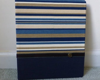 May include: A rectangular storage box with a navy blue bottom and a striped top. The stripes are in shades of blue, tan, and white. A dark brown band with a decorative button accents the front.