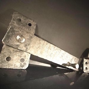 May include: A weathered, silver-toned metal gate latch. The latch has a rectangular design with multiple holes and a pivoting arm. The metal has a textured surface and shows signs of age.