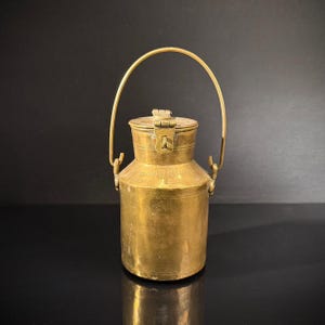May include: A vintage brass milk can with a hinged lid and a curved handle. The can has a cylindrical body with a slightly wider base and a narrower top. The handle is attached to the top of the can with two small brackets. The can is made of brass and has a shiny, golden finish.