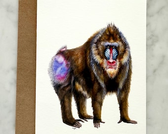 May include: A watercolor illustration of a mandrill primate, featuring brown fur, a blue and red face, and a purple and blue rear. The artwork is on a white card, accompanied by a brown envelope, suitable for greeting cards or art prints.