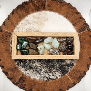 May include: A wooden box filled with various polished stones, including green, brown, and white stones. The box is sitting on a brown and white cowhide rug.