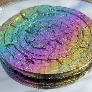 May include: A circular, iridescent, multi-colored, metallic object with a raised, textured design. The object has a rainbow sheen with shades of green, blue, purple, and gold.