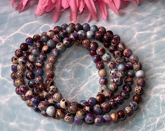 May include: Four strands of gemstone beads in shades of purple, brown, and blue are arranged in a spiral. The beads have a speckled pattern. A large pink flower is in the background, with a light blue water-like pattern.