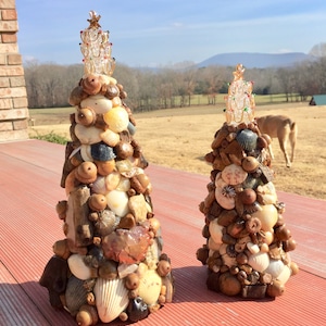 May include: Two miniature Christmas trees made of seashells, acorns, and driftwood. The trees are decorated with gold and red glass beads. The trees are on a red wooden surface.
