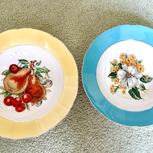 May include: Two decorative plates with scalloped edges. One plate is yellow with a pear and cherry design. The other is blue with a floral design. Both plates have a white center and gold trim.
