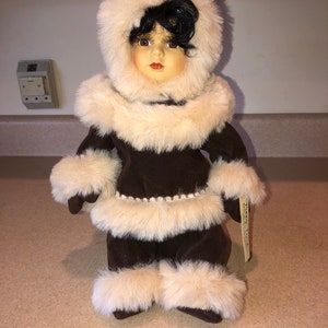 May include: A porcelain doll wearing a brown faux fur coat with a white fur trim. The doll has black hair and brown eyes. The doll is wearing a white beaded belt and has a tag that says "The Memory Tree".
