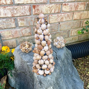 May include: A small, cone-shaped Christmas tree made of natural materials, such as nutshells and pinecones, decorated with white beads. The tree is sitting on a large, gray rock.