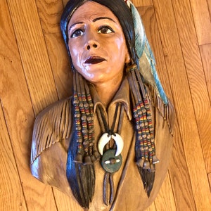 May include: A carved wooden bust of a Native American woman. The sculpture features detailed facial features, long dark hair, and traditional adornments, including a feather and beaded braids. The bust is painted in natural tones.