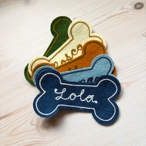 Op de afbeelding: Een stapel vilten hondenpenningen in de vorm van een bot in verschillende kleuren, waaronder marineblauw, lichtblauw, oranje, crème en groen. De marineblauwe tag heeft de naam "Lola" in het wit geborduurd.