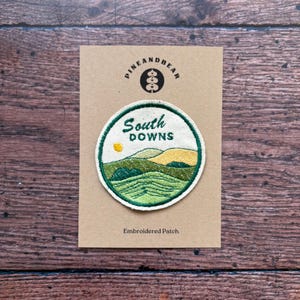 May include: Circular embroidered patch with a landscape design and the words "South Downs." The patch has a green and white border. The background is tan, with the brand name "PINEANDBEAR" at the top. The text "Embroidered Patch" is at the bottom.