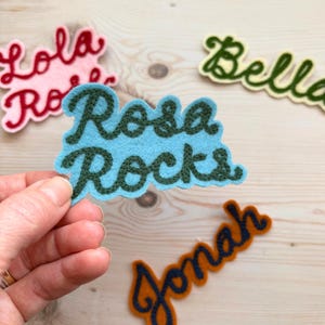 Pode incluir: Um patch de feltro azul com as palavras "Rosa Rocks" bordadas em verde escuro é segurado por uma mão. Outros patches de feltro com nomes como "Lola", "Bella" e "Jonah" estão espalhados. Os patches são em várias cores, incluindo rosa, verde e laranja.
