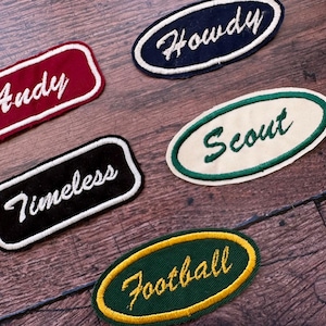 Op de afbeelding: Een verzameling geborduurde patches in verschillende vormen en kleuren. De patches bevatten de woorden "Andy", "Howdy", "Scout", "Timeless" en "Football". De patches zijn in rood, marineblauw, zwart, groen en geel.