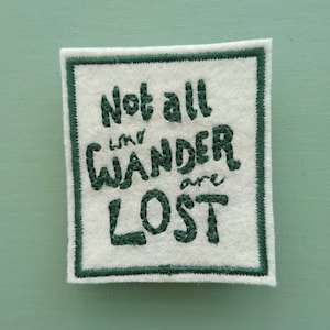 Puede incluir: Parche de tela blanca con texto bordado en verde que dice "Not all who wander are lost".
